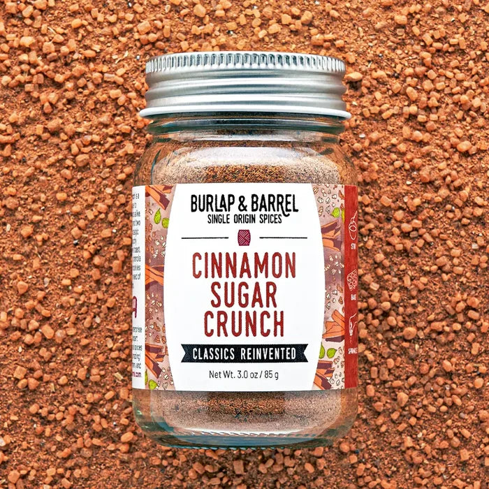 A glass jar of Burl & Barrel Cinnamon Sugar Crunch spice mix, featuring a white label with red and black text and a silver metal lid, set against a background of cinnamon bark pieces.