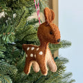 A brown felt deer ornament with white spots hanging from a red-and-white string on a green Christmas tree.